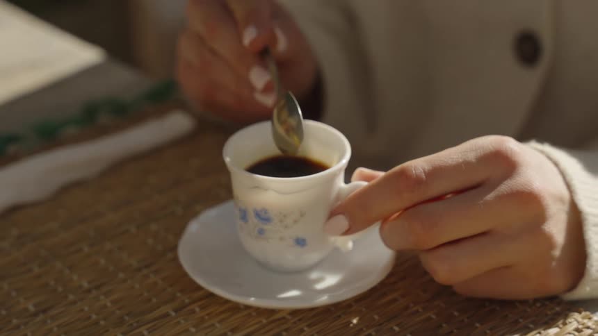 A woman is using a spoon to stir coffee in a dainty coffee cup