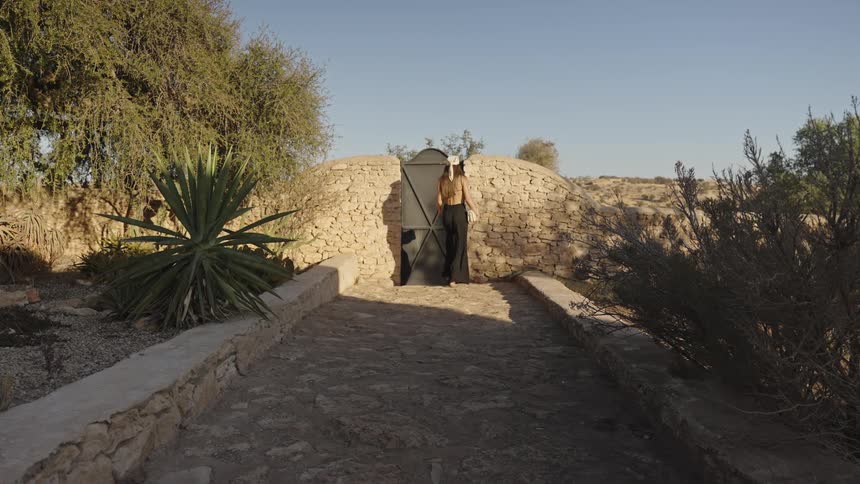 Young woman wearing a black dress and a white bandanna walking down a cobblestone walkway to a gate and then opening and crossing it outdoors during day time