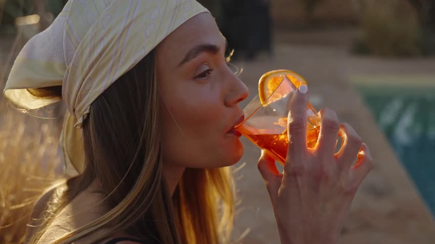 A close view shows a woman wearing a head scarf sitting poolside and sipping a bright orange cocktail garnished with an orange slice