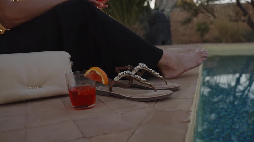 A closeup shot captures the legs and bare feet of a woman sitting by the edge of a swimming pool next to a glass of a red cocktail and a pair of sandals as the woman enjoys a moment of rest