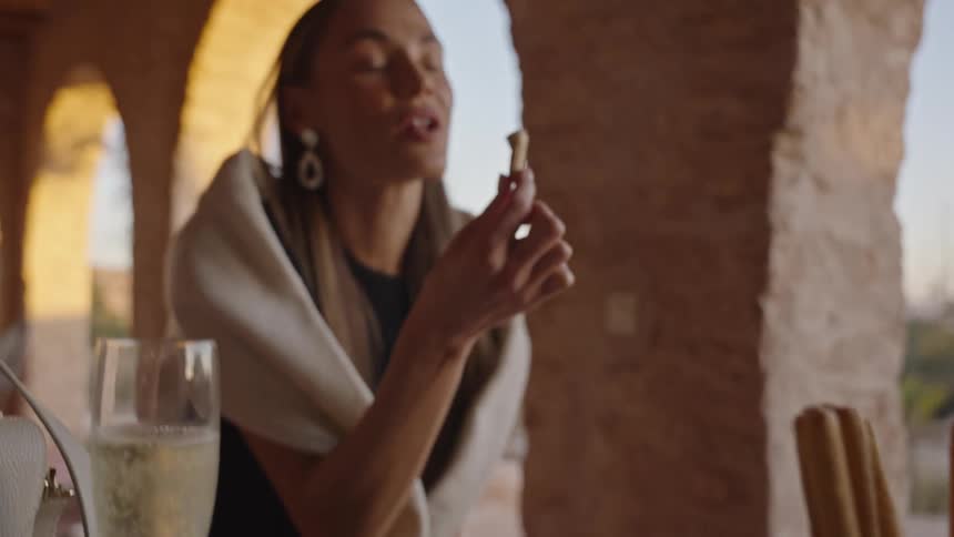 A woman is enjoying an outdoor meal or appetizer on a patio with stone arches visible in the background likely in a warm climateshe is dipping a breadstick into a small bowl of food with a glass of champagne or sparkling wine and a bowl of olives or