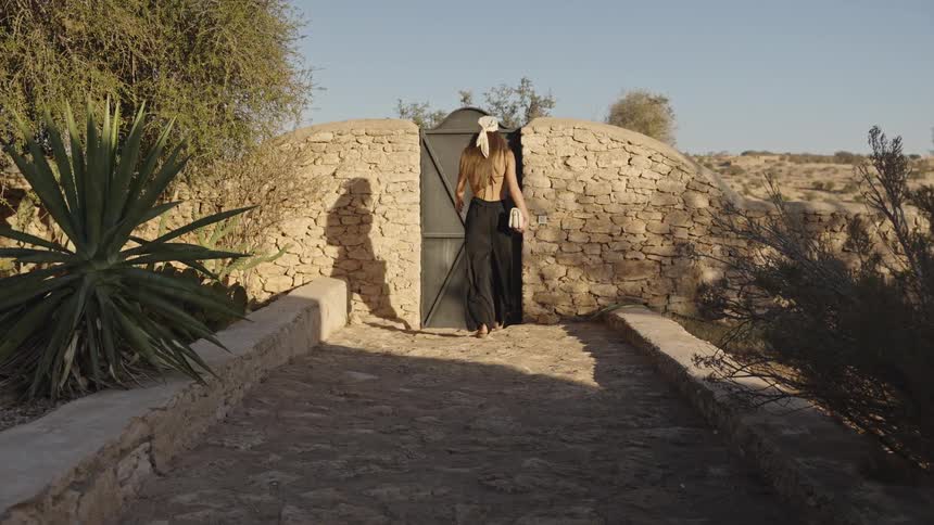 A woman wearing a black backless top and scarf walks down a stone path and opens a black metal door set in a rounded stone wall in an arid landscape