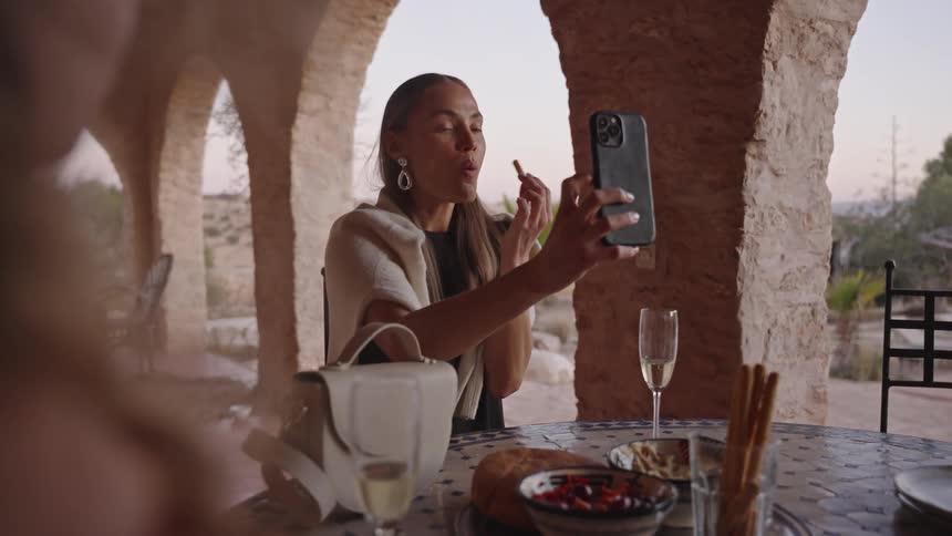 The video shows a woman seated outdoors under an arched stone patio applying lipstick while looking at her reflection in her smartphone she is at a table with a friend where glasses of champagne and snacks are laid out in a sunny rustic setting