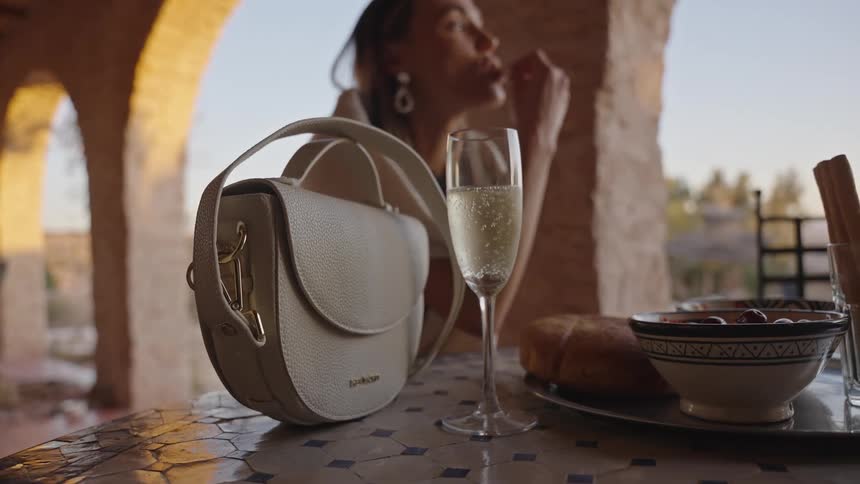 A closeup shot focuses on a purse and a bubbling glass of sparkling wine on an outdoor patio table with small bowls of food nearby and a woman enjoying an appetizer in the softly blurred background against an arched sunlit architectural setting