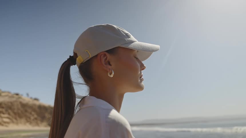 A young woman in a cap stands peacefully in profile against a sunny coastal backdrop