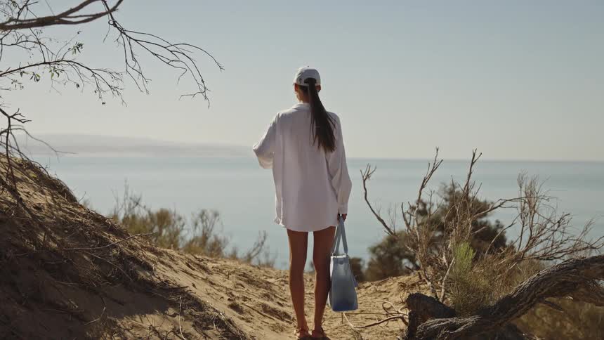 A woman stands on a sandy dune overlooking the bright blue ocean