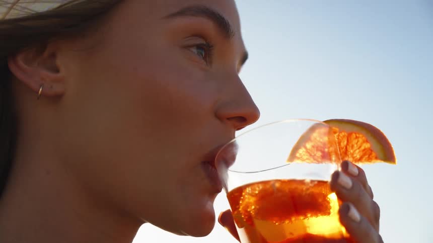 The face of a blonde woman drinking from a glass containing ambercolored liquid