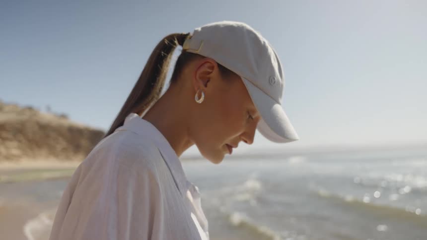 A close side view of a woman with a ponytail and baseball cap standing on a sunny beach looking down toward the glittering water