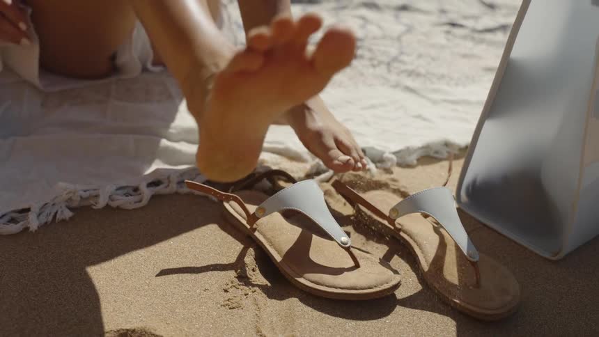 A woman sitting on a patterned beach blanket on the sand adjusting the strap of a sandal on their foot before relaxing her legs with a white shopping bag visible nearby