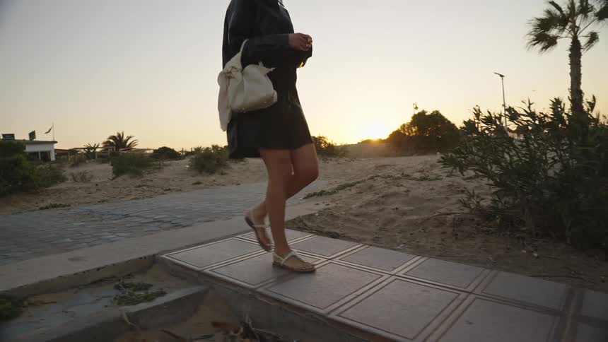 The video shows a woman with long dark hair wearing a black outfit and lightcolored sandals walking along a paved walkway near a sandy outdoor area the scene is set around sunset with a bright orange glow visible over the lowlying buildings and veg