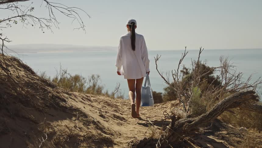 A young woman in a white longsleeve shirt holding a handbag walks toward the beach on a sunny day