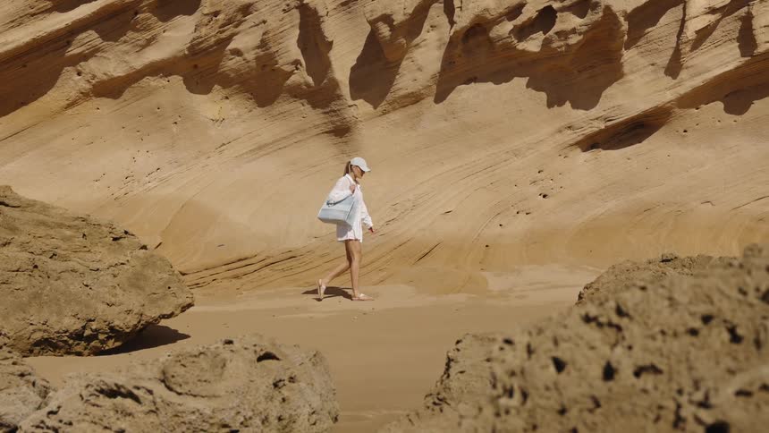 A wide view shows a woman wearing a white cap and sandals walking between large foreground rocks and steep tan sandstone cliffs