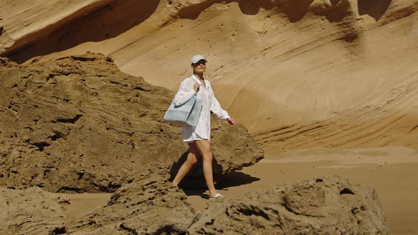 A close view shows a woman wearing a white cap and tunic walking across a rocky sandy beach near huge layered orange cliffs