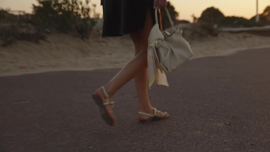 Woman walks with lightcolored bag on beachside path during sunset