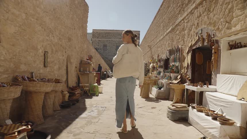 A camera follows behind a woman with a tote bag as she walks slowly through a narrow sunlit alleyway lined with rough stone walls and various stalls selling local handmade goods and crafts