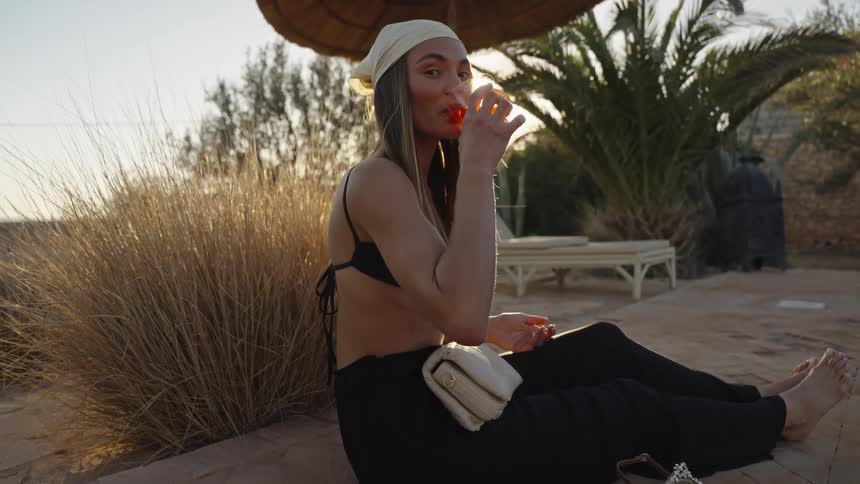A young woman wearing a white headscarf a black bikini top and black trousers is sitting barefoot outdoors on a stone surface taking a sip from a glass containing a cocktail with strong sunlight palm trees dry grass and a thatched umbrella visible