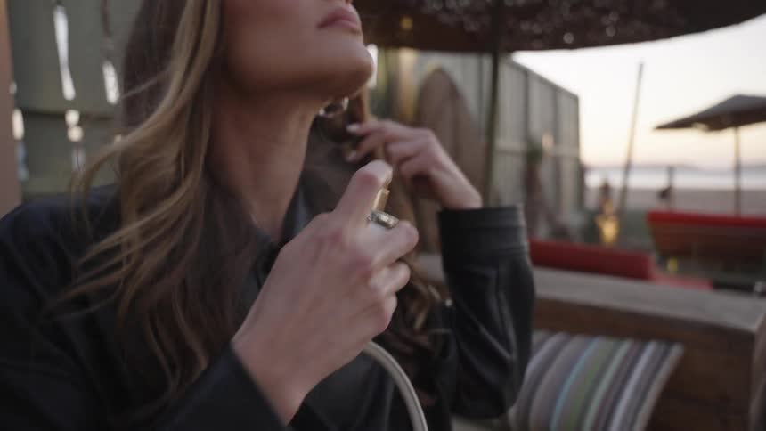 Woman with wavy hair holding spray bottle in outdoor setting with striped cushions at sunset