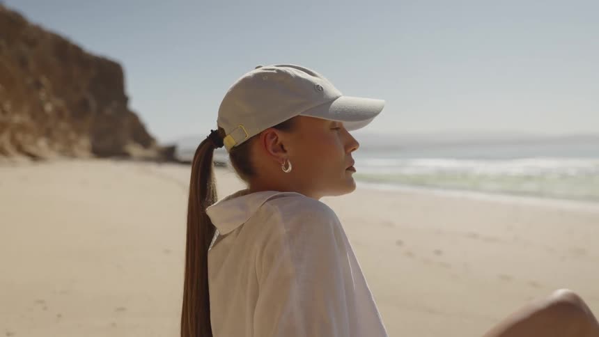 Woman sits on sandy beach gazing at ocean under clear sky and bright sun in quiet reflection
