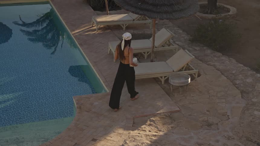 A woman walking along the stone path next to a swimming pool holding a drink and a small bag before stopping to place the drink on a side table next to a lounge chair under a thatched umbrella