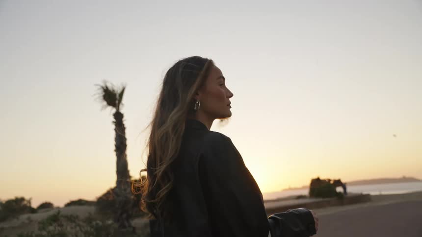 A woman in a dark dress is looking forward while walking on an asphalt path and carrying a lightcolored handbag at sunset witch a beach in the background