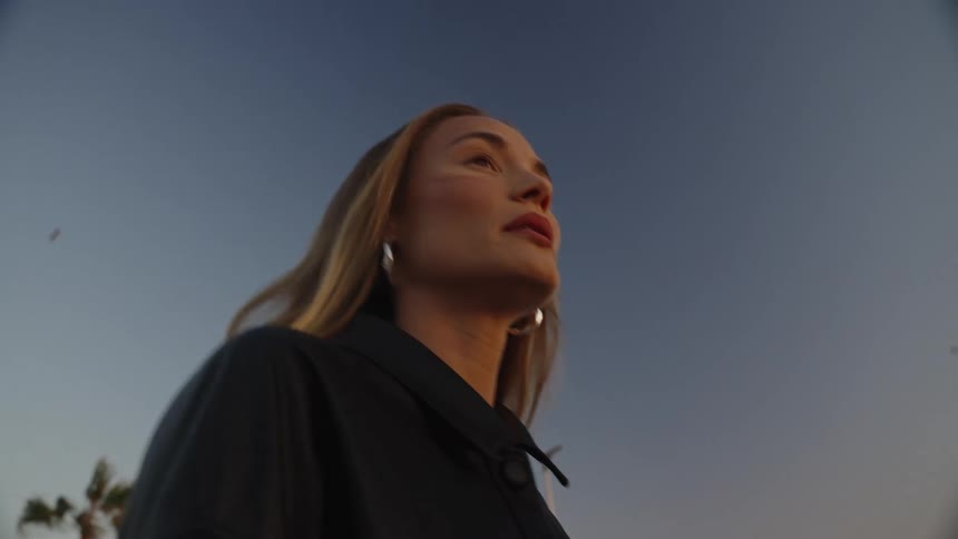 A young woman with blonde hair wearing round silver earrings and a black dress with makeup on her face walks outdoors at sunset