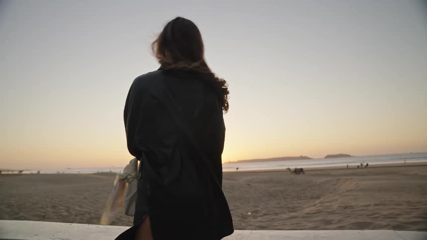A woman in a dark outfit possibly a leather jacket or jumpsuit is filmed closeup outdoors near a beach with the sunset visible over the ocean in the backgroundshe turns away from the camera and then quickly turns back towards it letting her hair f