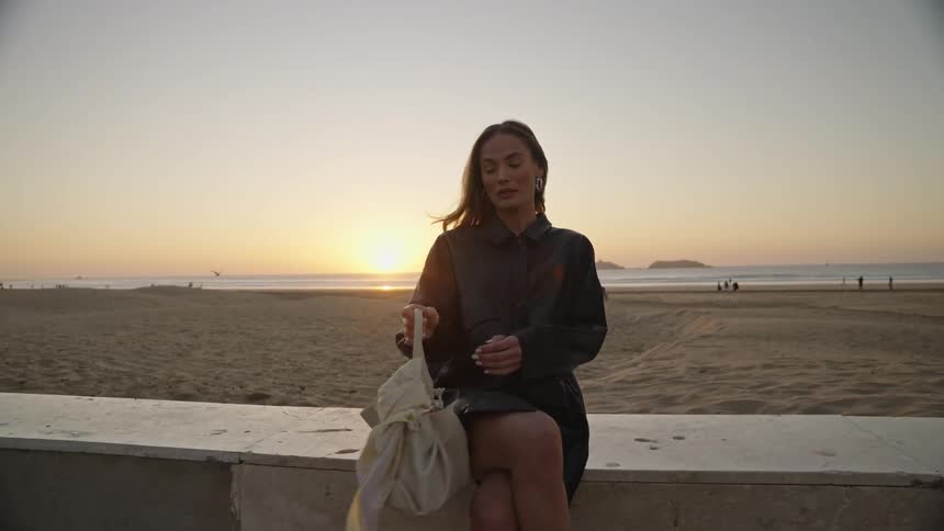A woman in a dark shirtdress or jacket sits on a low concrete wall overlooking a wide sandy beach the video is filmed during sunset casting an orange glow over the sceneshe is holding a lightcolored drawstring bag and adjusts a silk scarf tied to
