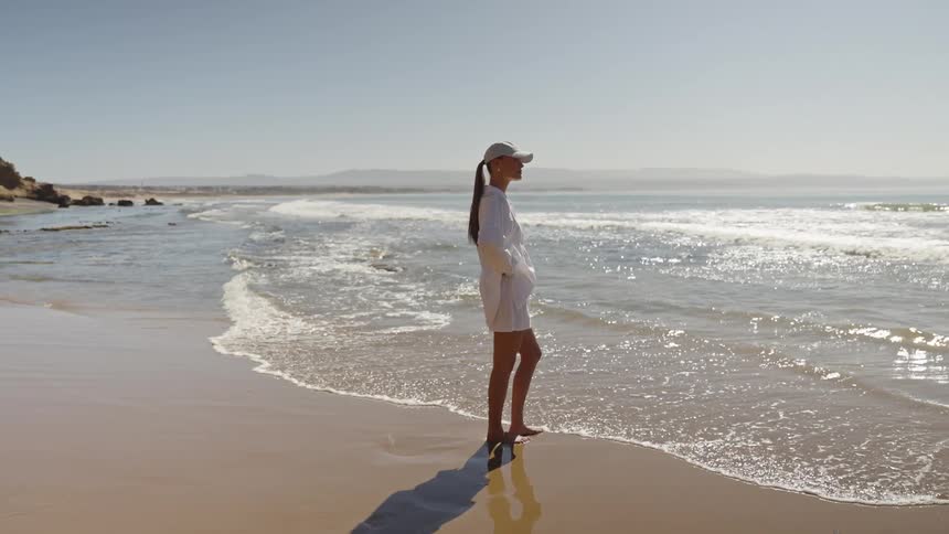 A woman wearing all white and a baseball cap stands barefoot on a sunny beach gazing out at the ocean waves gently rolling in the shot is a steady side profile showing the vast coastline and the womans shadow extending onto the wet sand