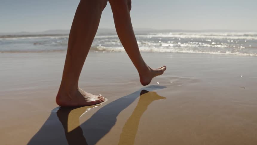 A close view shows a person walking barefoot across the firm wet sand casting a long shadow with the sparkling ocean waves in the distance