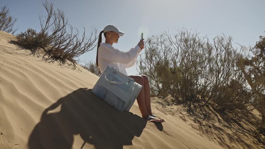 A woman sitting on the steep slope of a large sand dune using her mobile phone