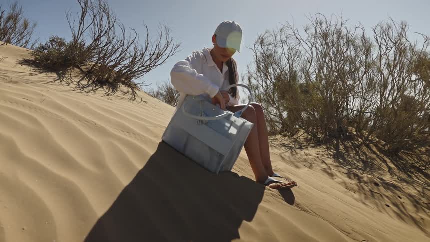 A woman walking up a large sand dune then sitting down in the bright sun where she pulls out her phone from a large tote bag and begins to look at the screen