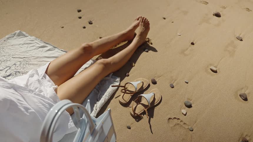 The bare legs and feet of a woman lying down on a white blanket on a sandy beach with a pair of brown and light blue sandals a small beach bag and scattered footprints and pebbles visible on the sunlit sand nearby