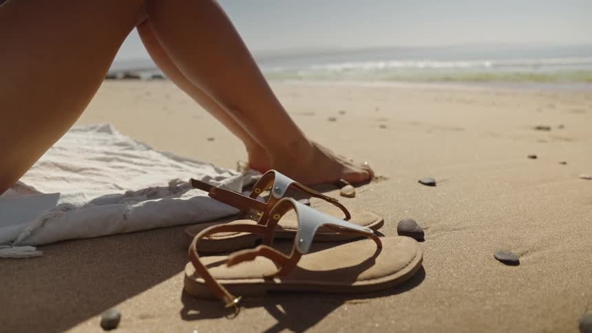 A closeup shot captures a pair of light blue and tan sandals lying on a sandy rocky beach next to a persons bare feet and legs the person is sitting on a white towel with the ocean waves and coastline visible suggesting a sunny day by the sea