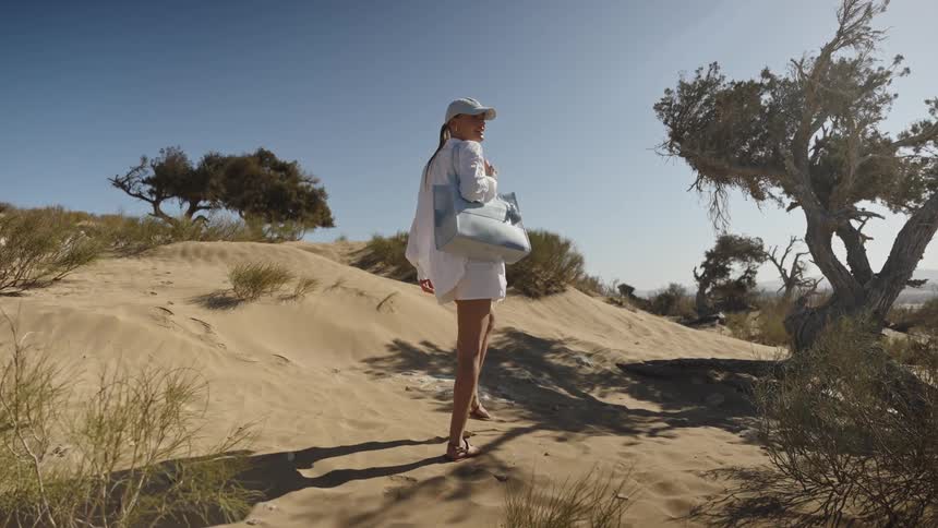 A woman carrying a bag on her shoulder or walking on a sandy desert area with some lowlying vegetation