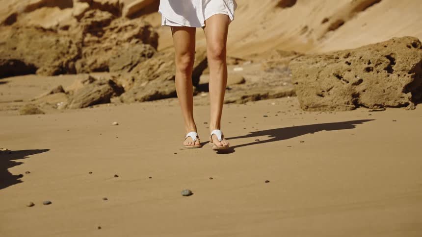 A stylish woman in a white oversized shirt sandals and a cap walks on a sunlit sandy beach with tall rocky cliffs in the background