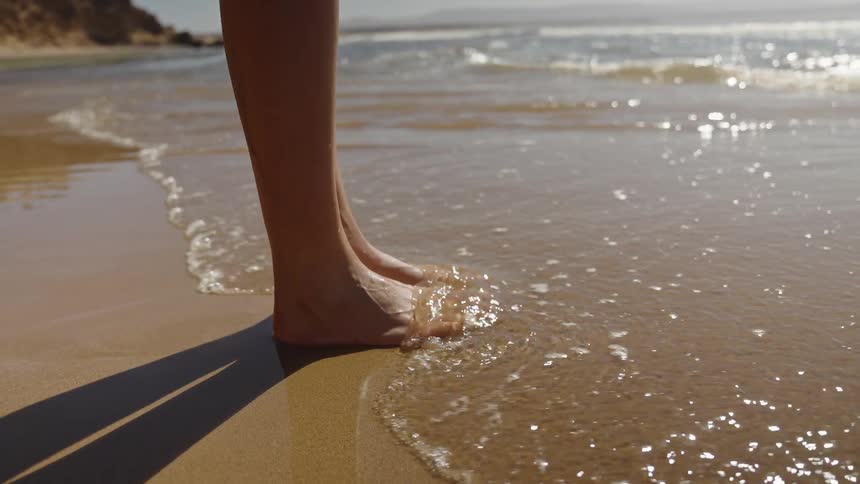 A close view shows bare feet standing on wet beach sand as a small sparkling wave gently washes over them