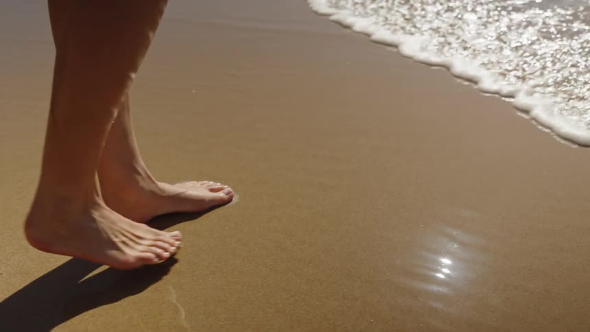 Woman walks barefoot on the beach as waves roll into shore and over her feet