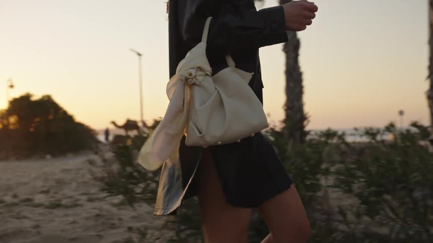 A longhaired woman wearing a dark coat or dress and carrying a small lightcolored backpack on her shoulder walks past palm trees during sunset with a silhouette of camels visible in the distance