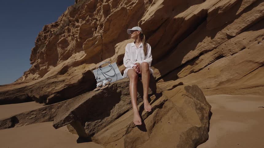 Person sitting barefoot on beachside rock with rugged cliff in the background