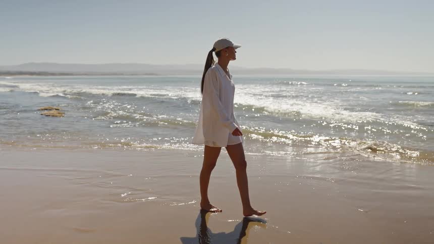 A woman walks barefoot on wet sand as waves roll in on a sunny beach with distant cliffs