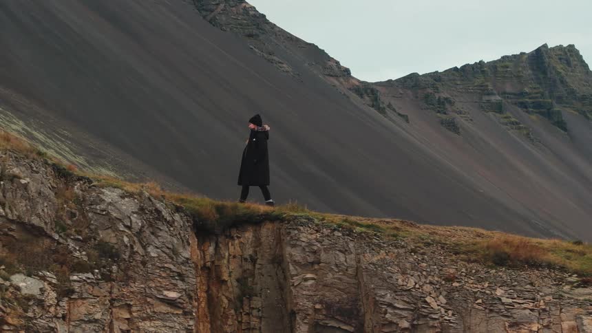 A lone figure dressed in all black stands near the edge of a rocky cliff overlooking a sheer drop the background is dominated by a massive dark and rugged mountain face suggesting a dramatic desolate landscape