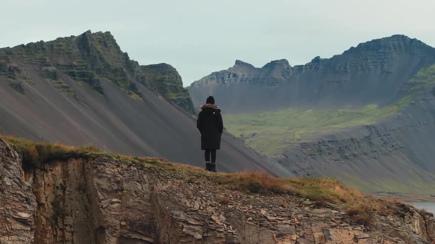 A lone figure stands on a rocky cliff edge overlooking a majestic range of jagged mountains
