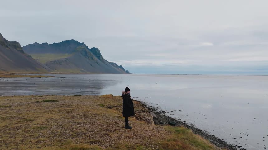 A solitary figure stands on a grassy cliff overlooking a coastal landscape with distant mountains