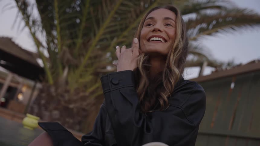 A closeup shot of a young woman sitting outdoors in the evening with a palm tree in the background as she smiles and laughs genuinely bringing her hand up to touch her face