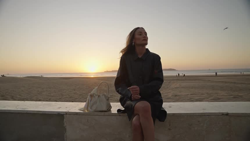 A young woman sits on a wall overlooking the beach at sunset