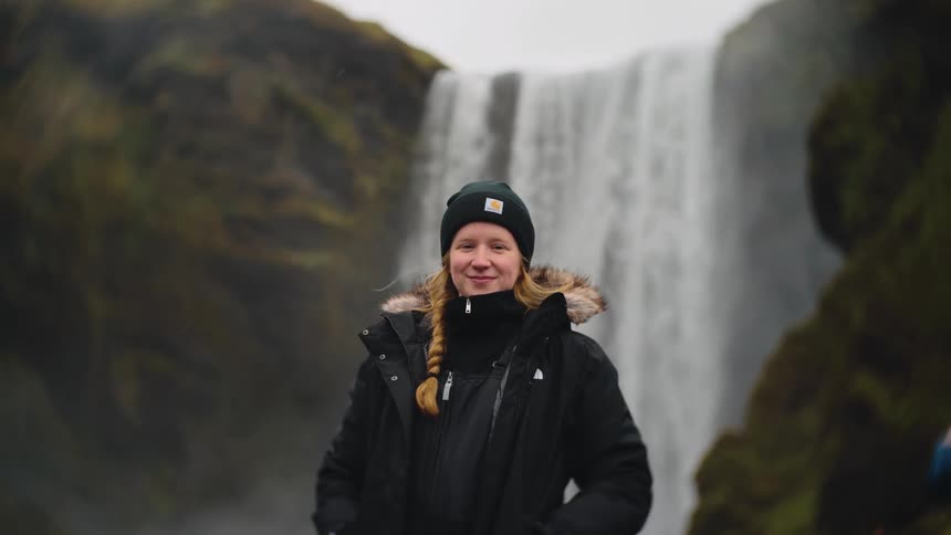 Young woman wearing a black jacket and a beanie standing next to a waterfall outdoors during day time