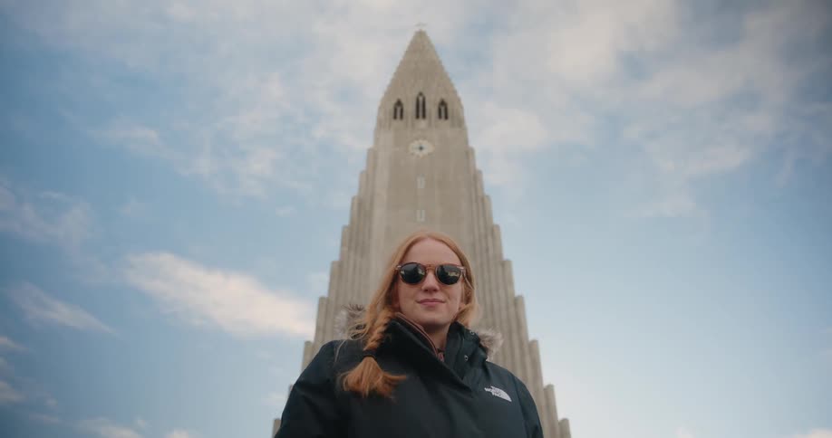 A smiling woman stands in front of a light colored church under a cloudy blue sky