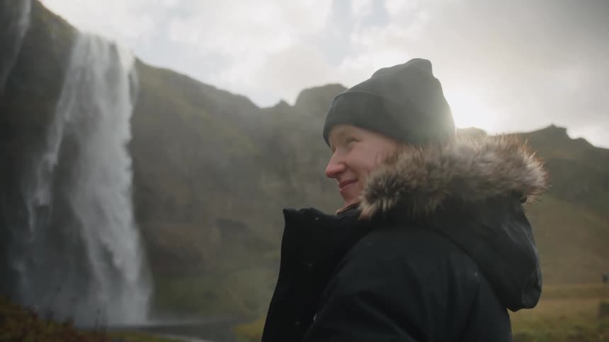 A close view shows a woman wearing a beanie and a fur trimmed parka smiling while observing a huge misty waterfall in a cold rugged landscape