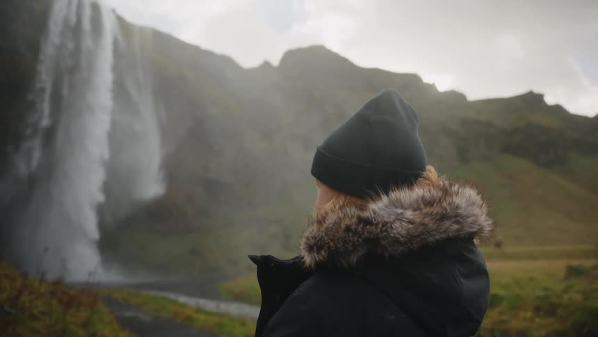 A woman wearing a dark coat with a fur hood and a beanie stands on a path and looks towards a majestic waterfall descending from high cliffs