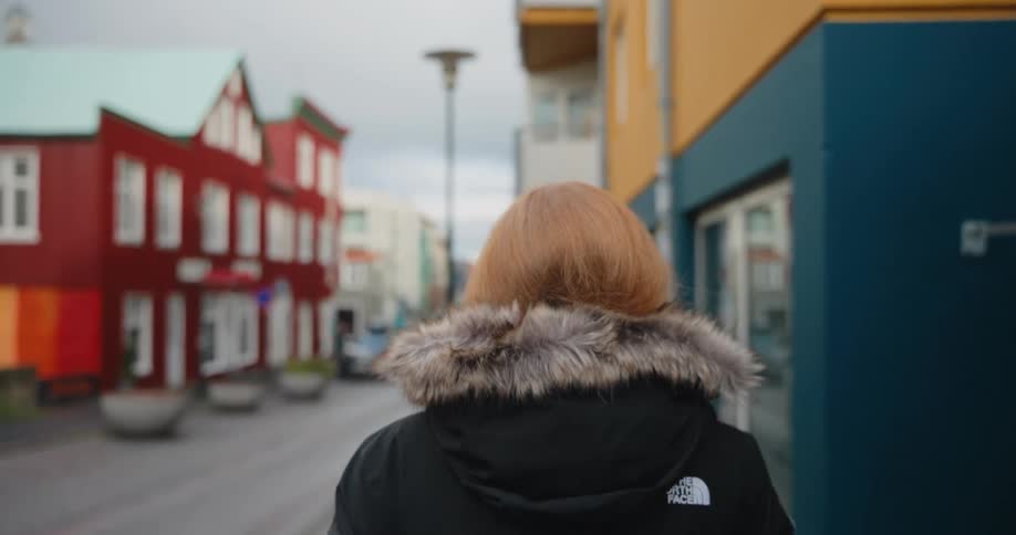 A close view from behind shows a woman wearing a black winter jacket with a fur hood walking along a street past bright colored buildings